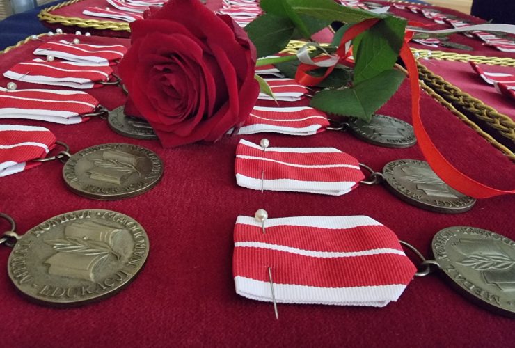 Na tacy pokrytej czerwoną tkaniną leżą medale Komisji Edukacji Narodowej. Na medalach leży czerwony kwiat.